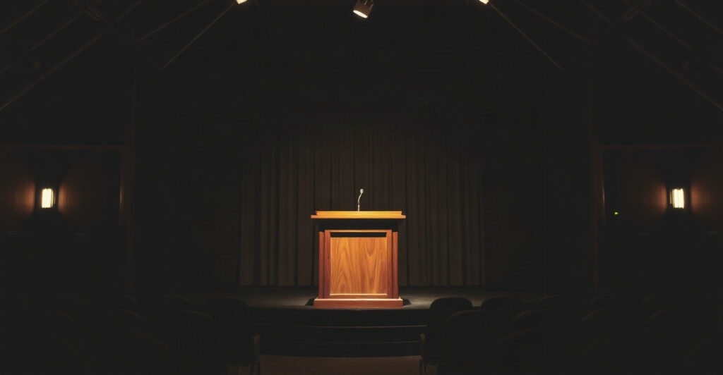 A lone lit lectern in a quiet hall, evoking the decades of lectures given by Neville Goddard